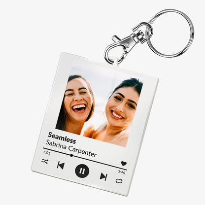 A custom acrylic keyring designed to resemble a digital music player interface. It features a square frame with a photo of two smiling people at the top. Below the photo are mock song details, including placeholder text 'Song Title Here' and 'Artist Name'. Music control icons—rewind, play, forward, and a heart—are displayed along the bottom, mimicking a streaming app. The keyring is attached to a metal clasp and keyring loop.