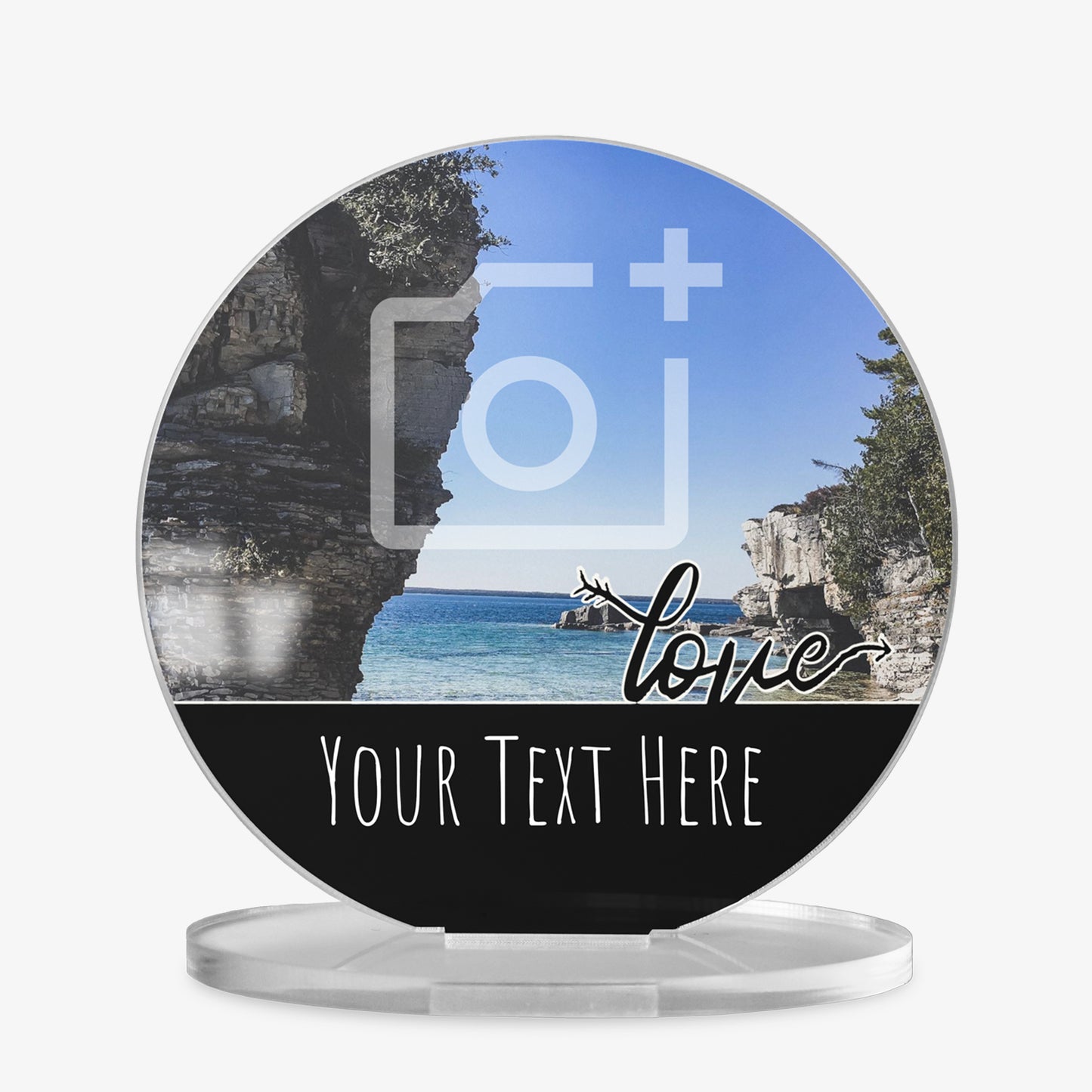A round acrylic display piece with a scenic coastal photo showing cliffs and blue sea under a clear sky. A placeholder camera icon is overlaid on the image, showing a customisable photo area. The bottom section has a black band with the word 'love' in cursive and space for personalised text reading 'Your Text Here'. The item is displayed on a clear round stand.