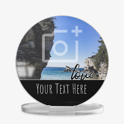 A round acrylic display piece with a scenic coastal photo showing cliffs and blue sea under a clear sky. A placeholder camera icon is overlaid on the image, showing a customisable photo area. The bottom section has a black band with the word 'love' in cursive and space for personalised text reading 'Your Text Here'. The item is displayed on a clear round stand.