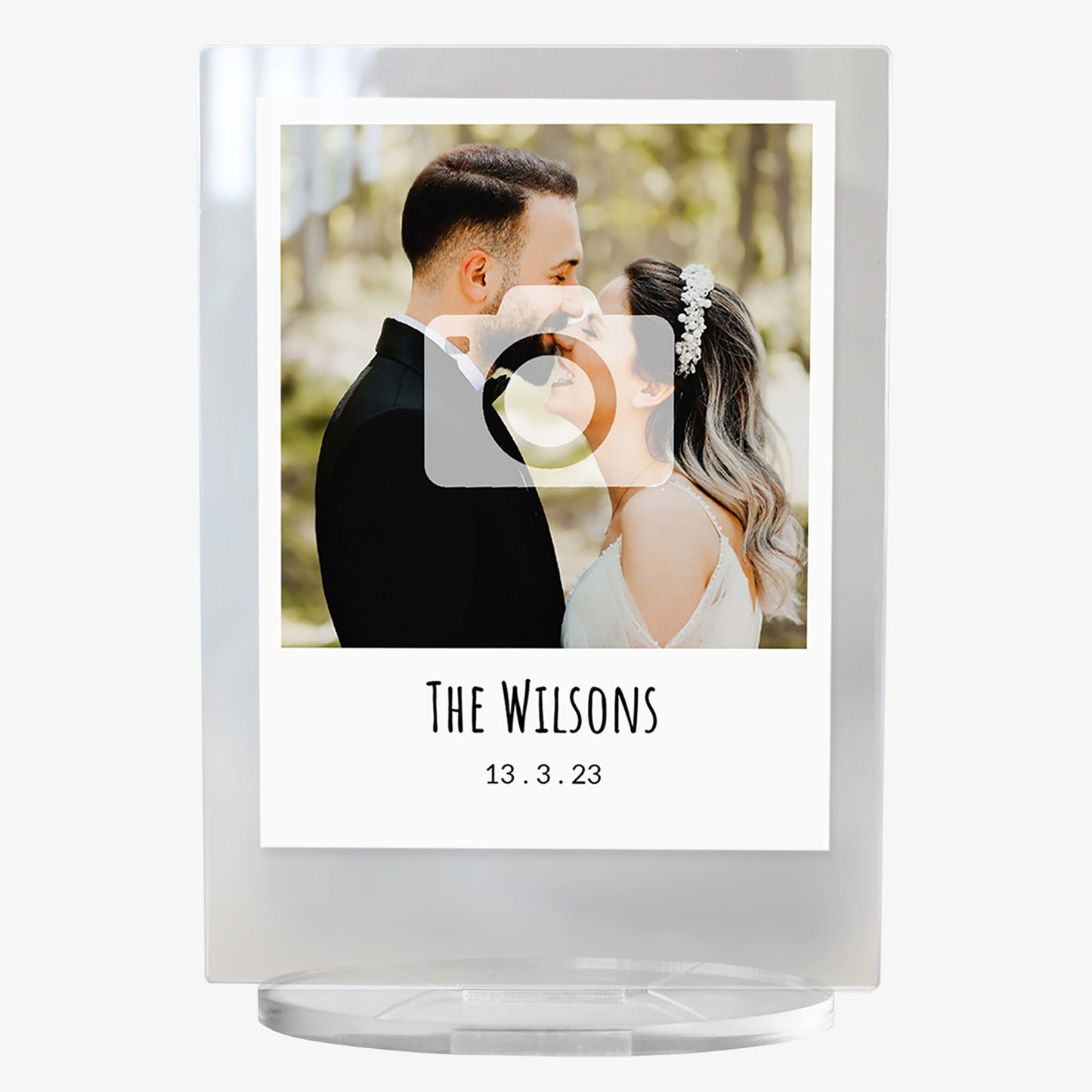 A personalised acrylic photo plaque with a clear stand. The plaque features a wedding photo of a bride and groom standing close together in a wooded setting. Below the image, text reads 'The Wilsons' followed by the date '13.3.23' in a simple, elegant font. The design mimics a polaroid-style frame for a modern keepsake look.