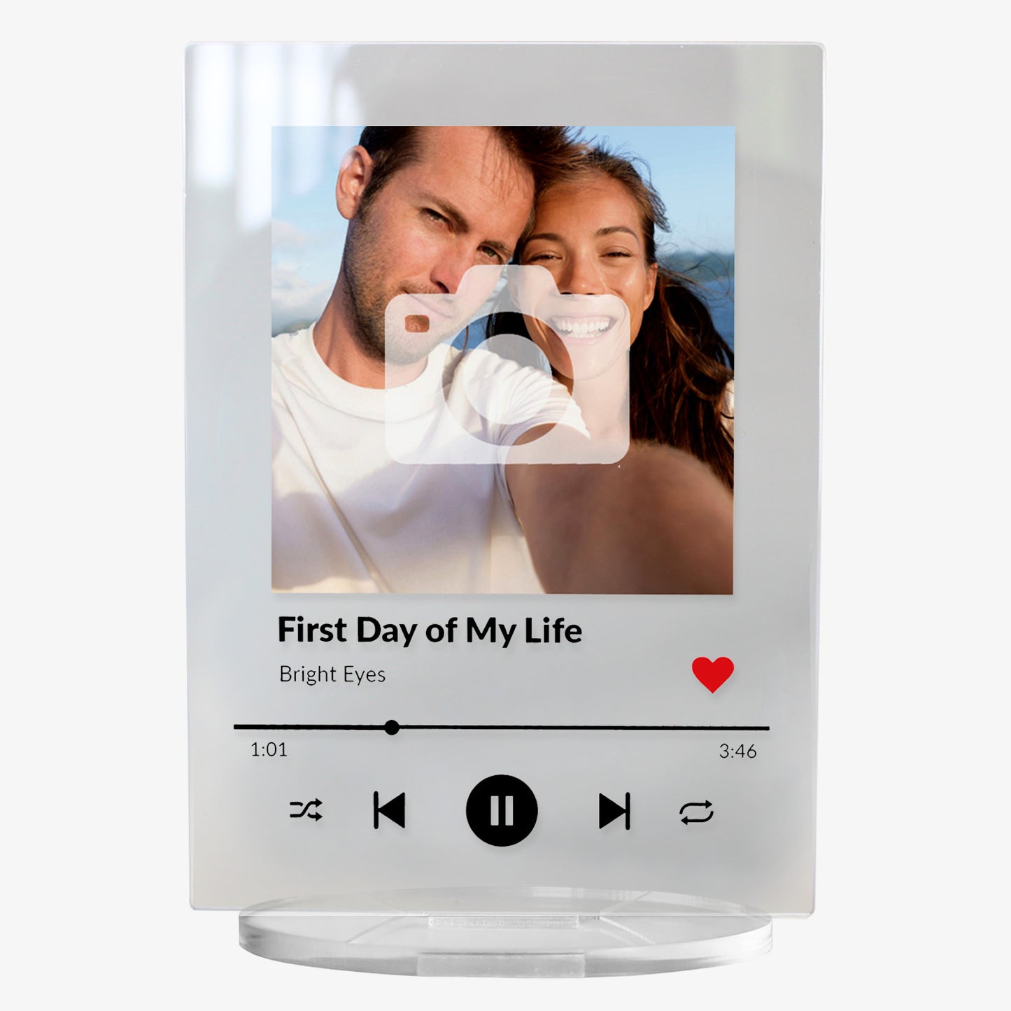 A personalised acrylic music plaque styled like a digital music player. It features a photo of a smiling couple at the top, followed by song details: 'First Day of My Life' by Bright Eyes. Music player controls, a progress bar, and a red heart icon appear below, mimicking a streaming app interface. The plaque stands upright on a clear circular base.