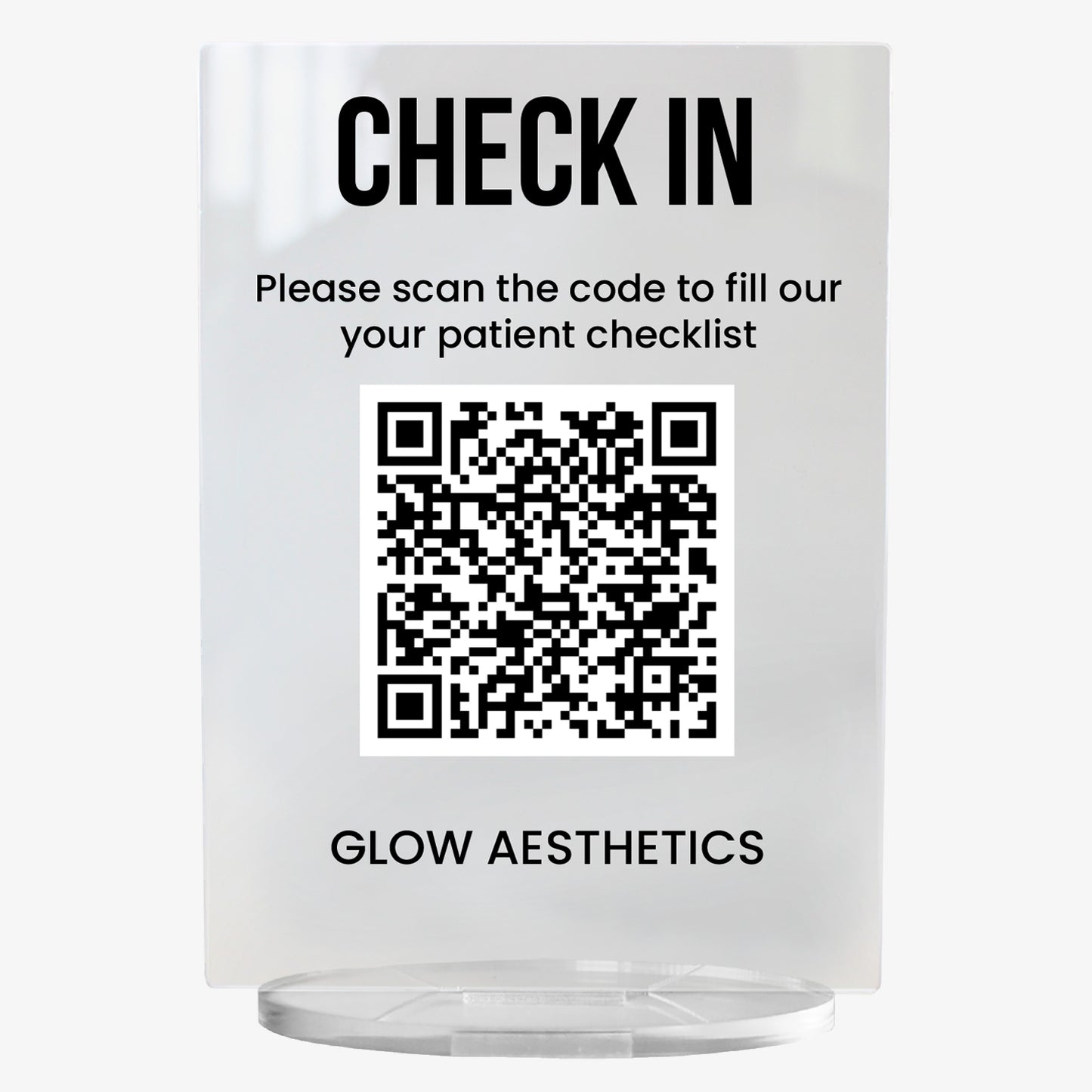 A clear acrylic sign with bold black text at the top reading 'CHECK IN'. Below, it says 'Please scan the code to fill out your patient checklist'. A large black and white QR code is centrally displayed, and the text 'GLOW AESTHETICS' appears at the bottom. The sign stands upright on a circular clear base.