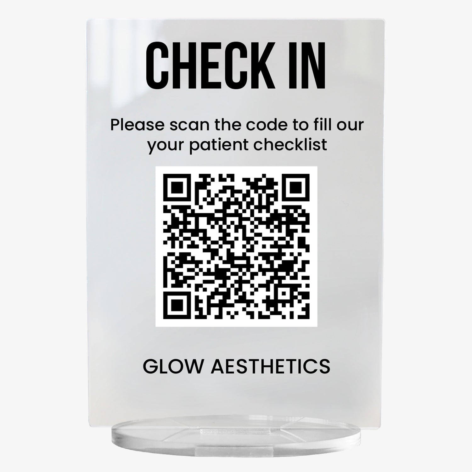 A clear acrylic sign with bold black text at the top reading 'CHECK IN'. Below, it says 'Please scan the code to fill out your patient checklist'. A large black and white QR code is centrally displayed, and the text 'GLOW AESTHETICS' appears at the bottom. The sign stands upright on a circular clear base.