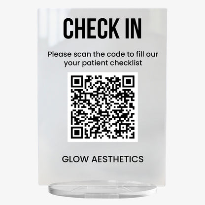A clear acrylic sign with bold black text at the top reading 'CHECK IN'. Below, it says 'Please scan the code to fill out your patient checklist'. A large black and white QR code is centrally displayed, and the text 'GLOW AESTHETICS' appears at the bottom. The sign stands upright on a circular clear base.