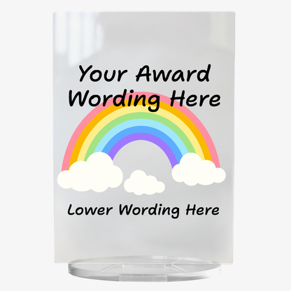 A clear acrylic award plaque standing on a circular base, featuring a large rainbow with either end in a cloud in the centre. Overlaying the rainbow is placeholder text reading 'Your Award Wording Here' in bold black font. Below, additional text reads 'Lower Wording Here'. 