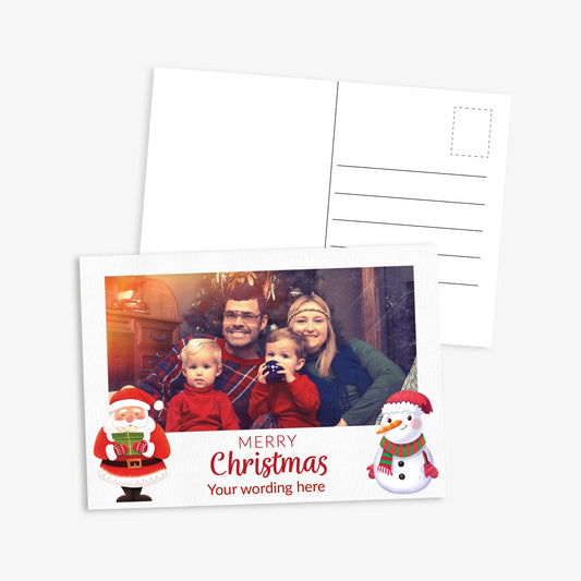Front and back of a festive Christmas postcard. The front features Father Christmas and a snowman either side of the text 'Merry Christmas' with placeholder below 'Your Wording Here' and space to upload own photo - the image shows a family in front of a christmas tree. The back includes standard postcard lines for address and message
