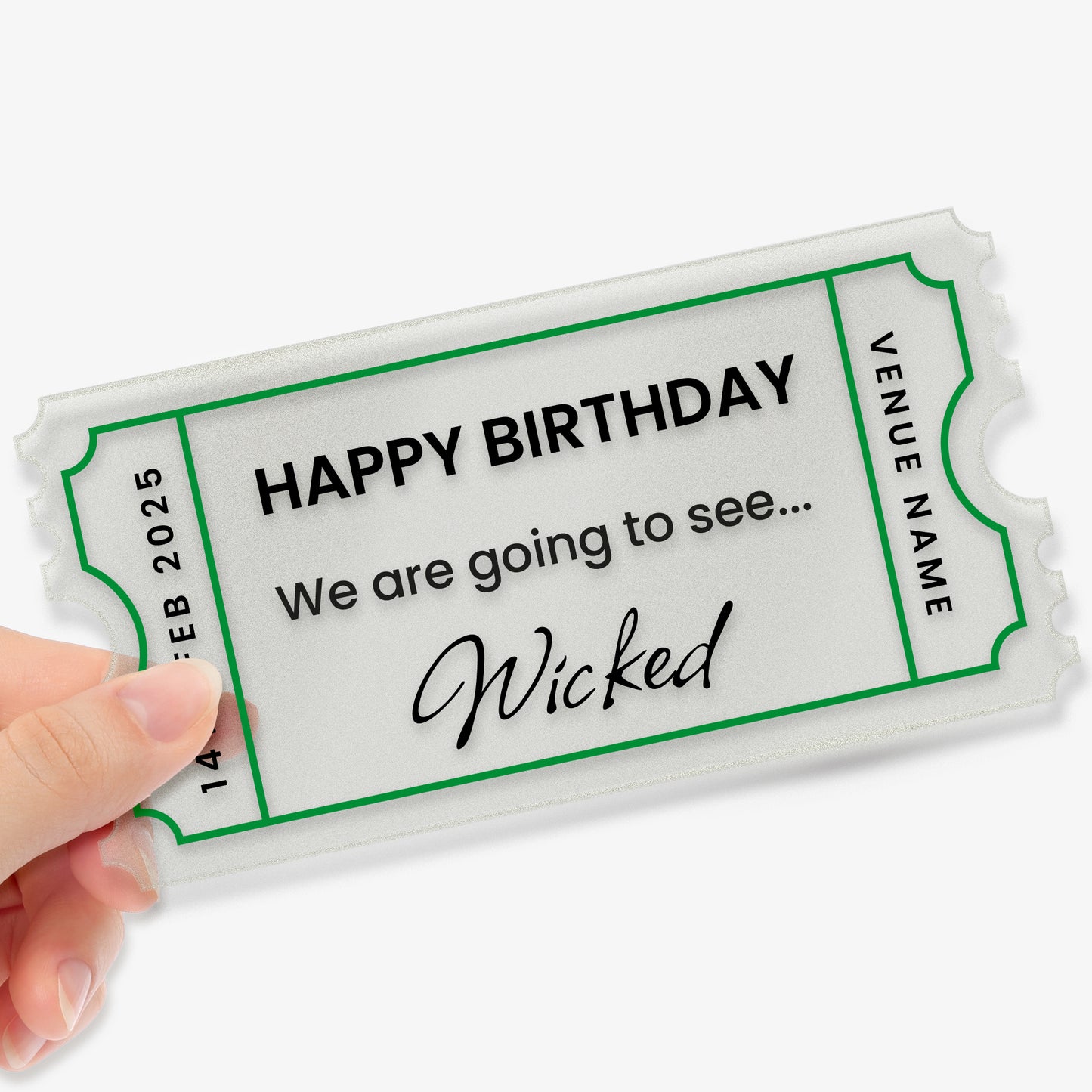 A hand holding a custom acrylic gift ticket styled like an event pass, with decorative notched edges and a green border. The text reads: 'HAPPY BIRTHDAY – We are going to see... Wicked'. The left side shows the date '14 FEB 2025', and the right side has a placeholder reading 'VENUE NAME'. The design mimics a theatre or concert ticket, making it a fun and memorable birthday gift.