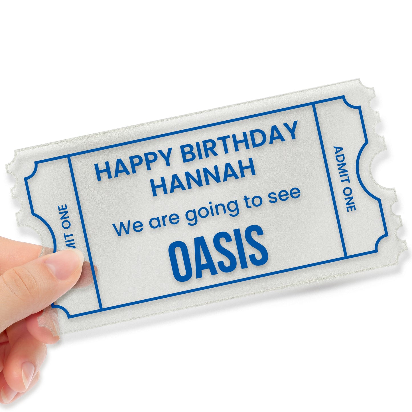 A hand holding a custom acrylic gift ticket styled like an event pass, with decorative notched edges and a blue border. The text reads: 'HAPPY BIRTHDAY HANNAH – We are going to see... OASIS'. The left side shows the placeholder 'ADMIT ONE', and the right side has a placeholder reading 'ADMIT ONEE'. The design mimics a theatre or concert ticket, making it a fun and memorable birthday gift.