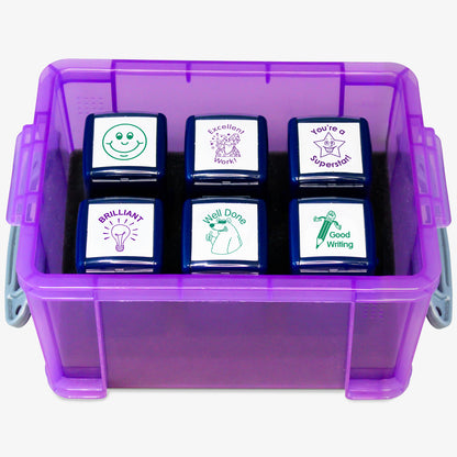 A transparent purple storage box containing six teacher stampers with positive feedback phrases and images. The stamps read: 'BRILLIANT!' with a light bulb, 'Well Done' with a smiling face, 'Good Work' with a pencil, 'Excellent Work' with a star character, 'You're a Superstar!' with a starburst, and a smiley face icon. The clear lid is slightly open, displaying the 'PTS Stampers' label.