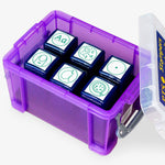 A transparent purple storage box containing six green ink teacher stampers with literacy-themed icons. The stamps display various symbols: uppercase and lowercase 'Aa', a face vocalising 'c-a-t', a magnifying glass with punctuation marks, a full stop, a traced lowercase 'a' for handwriting practice, and a vest shape representing personal effort or behaviour. The lid is open and labelled 'PTS Stampers'.