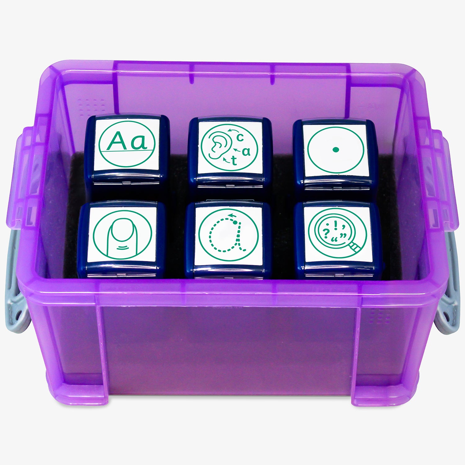 A transparent purple storage box containing six green ink teacher stampers with literacy-themed icons. The stamps display various symbols: uppercase and lowercase 'Aa', a face vocalising 'c-a-t', a magnifying glass with punctuation marks, a full stop, a traced lowercase 'a' for handwriting practice, and a vest shape representing personal effort or behaviour. The lid is open and labelled 'PTS Stampers'.