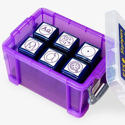 A transparent purple storage box containing six purple ink teacher stampers with literacy-themed icons. The stamps display various symbols: uppercase and lowercase 'Aa', a face vocalising 'c-a-t', a magnifying glass with punctuation marks, a full stop, a traced lowercase 'a' for handwriting practice, and a vest shape representing personal effort or behaviour. The lid is open and labelled 'PTS Stampers'.