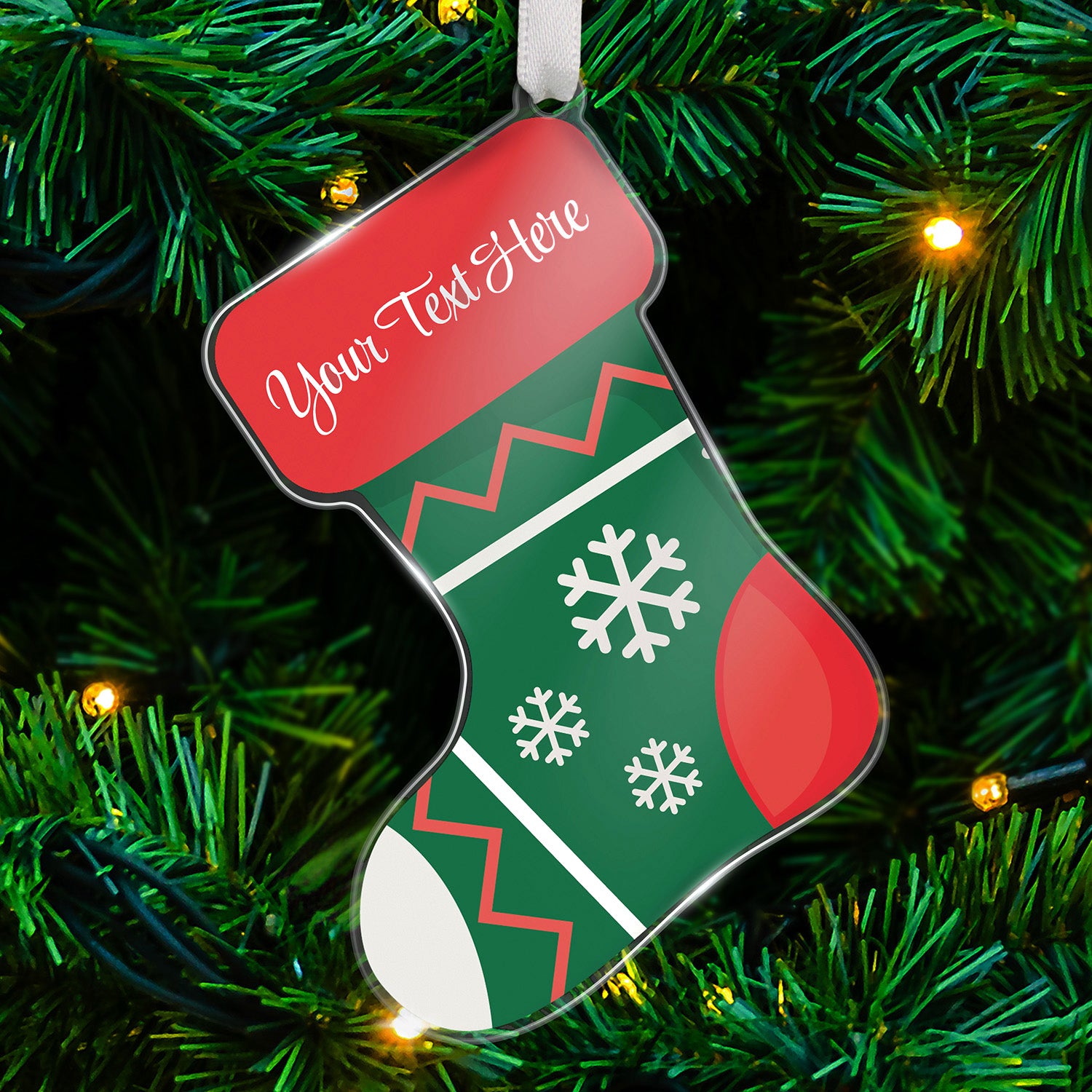 Personalised acrylic Christmas stocking-shaped tree ornament decorated with agreen background with white snowflakes and red zig zags on it; with a white toe of the stocking and red heel of the stocking. Featuring a red space at the top for customisation saying 'Your Text Here' in white script, hanging on a Christmas tree with lights.