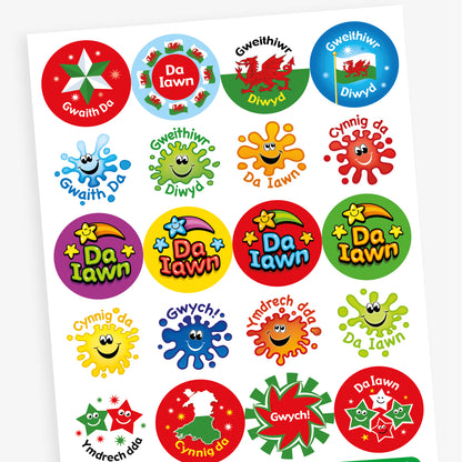 Overview of a sheet of Welsh-language reward stickers featuring various colourful designs, including a red Welsh dragon with 'Gweithiwr Diwyd', a roundel with Welsh flags and 'Da Iawn', a smiling paint splash with 'Gwaith Da', and a green germ character also labelled 'Gweithiwr Diwyd'.