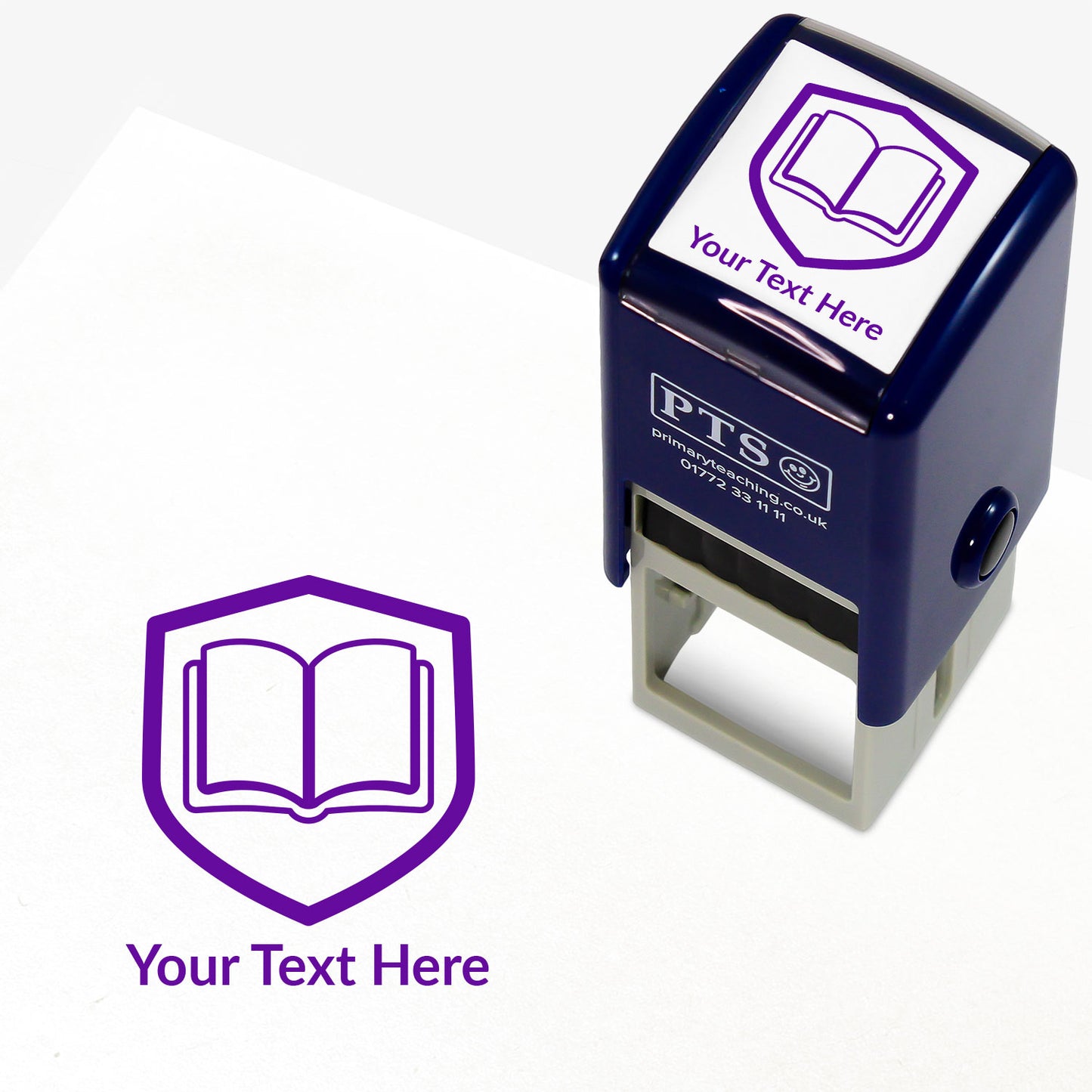 A stamper shown next to a sample stamped image on a white background. The stamp has a purple outline of an open book inside a shield shape, with placeholder text 'Your Text Here' on the top panel and below the stamp print.
