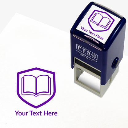 A stamper shown next to a sample stamped image on a white background. The stamp has a purple outline of an open book inside a shield shape, with placeholder text 'Your Text Here' on the top panel and below the stamp print.