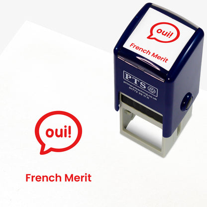 A stamper shown next to a sample stamped image on a white background. The stamp has a red outline of the word 'oui!' inside a shield shape, with placeholder text 'French Merit' on the top panel and below the stamp print.