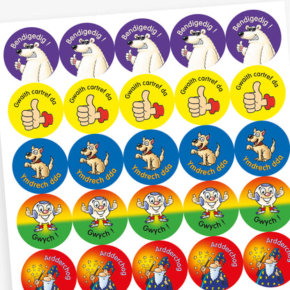 Sheet of circular stickers featuring cartoon characters and Welsh praise phrases. Includes: wizard with stars on a red background and the word 'Ardderchog'; A smiling tiger on a green background with the phrase 'Da iawn';  A thumbs-up hand on a yellow background with the phrase 'Gwaith cartref da'; A polar bear giving a thumbs up on a purple background with the word 'Bendigedig!'