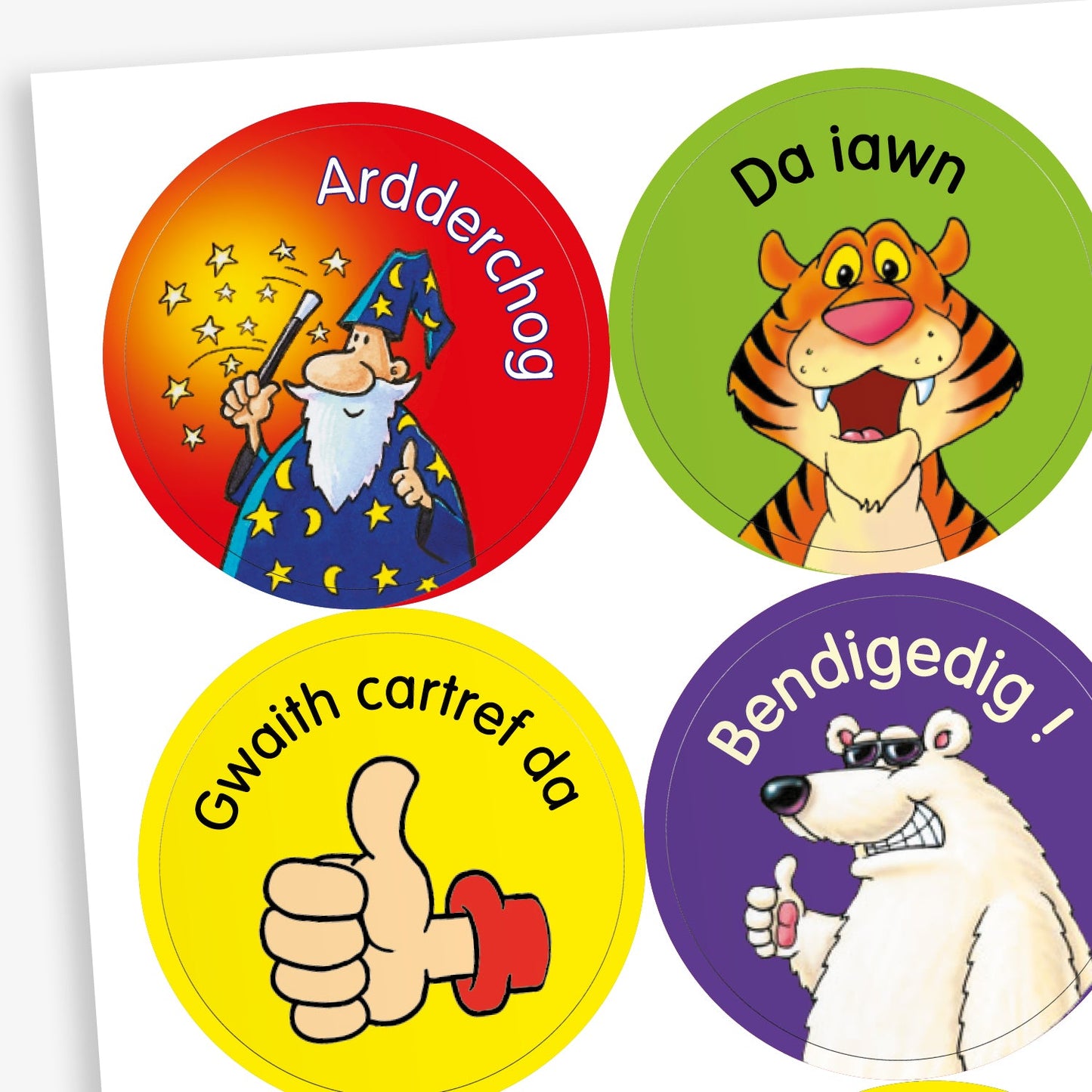 Sheet of circular stickers featuring cartoon characters and Welsh praise phrases. Includes: wizard with stars on a red background and the word 'Ardderchog'; A smiling tiger on a green background with the phrase 'Da iawn';  A thumbs-up hand on a yellow background with the phrase 'Gwaith cartref da'; A polar bear giving a thumbs up on a purple background with the word 'Bendigedig!'