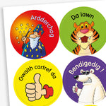 Sheet of circular stickers featuring cartoon characters and Welsh praise phrases. Includes: wizard with stars on a red background and the word 'Ardderchog'; A smiling tiger on a green background with the phrase 'Da iawn';  A thumbs-up hand on a yellow background with the phrase 'Gwaith cartref da'; A polar bear giving a thumbs up on a purple background with the word 'Bendigedig!'