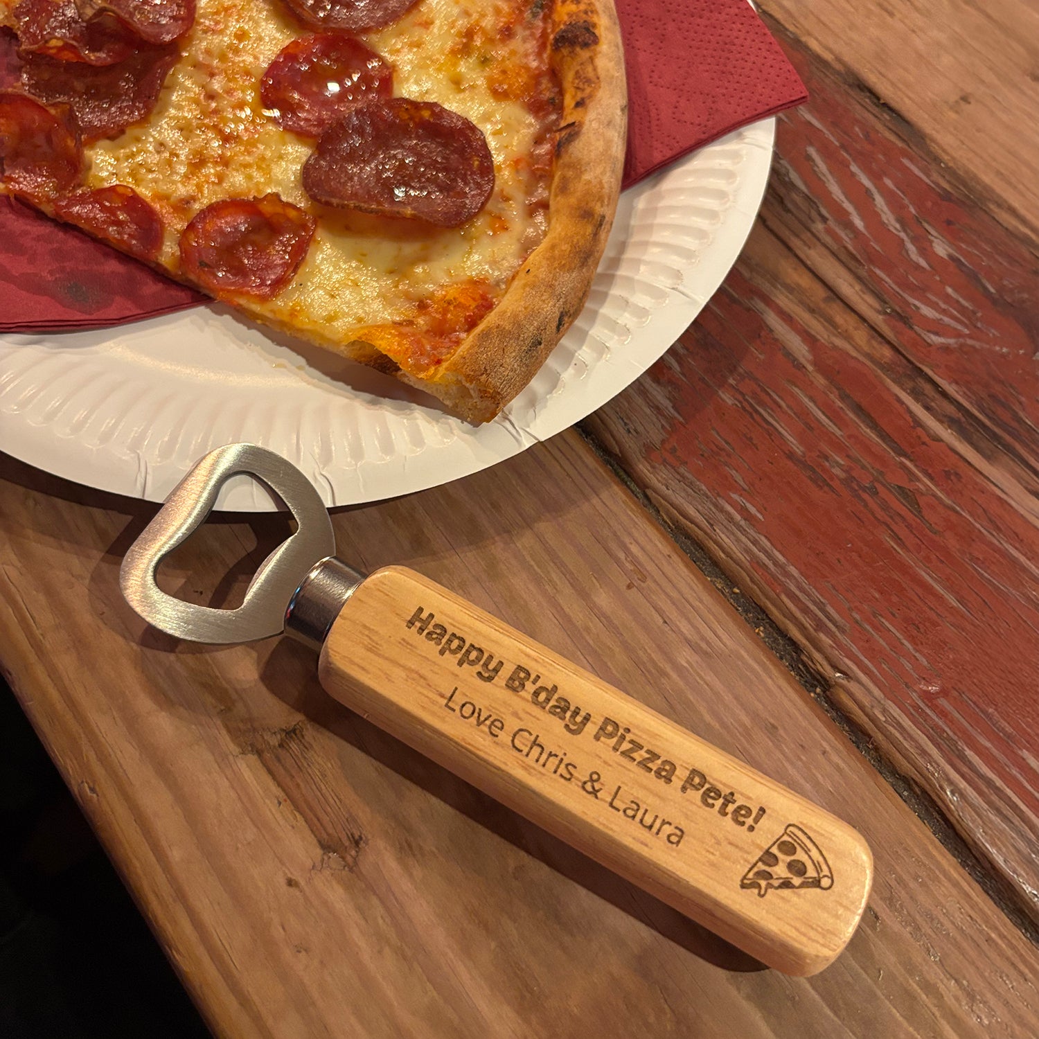 Personalised bottle opener on wooden table next to a plate of pepperoni pizza. Example text etched onto the wooden handle of the bottle opener reads "Happy B'day Pizza Pete! Love, Chris and Laura" with an image of a pizza slice etched to the right.