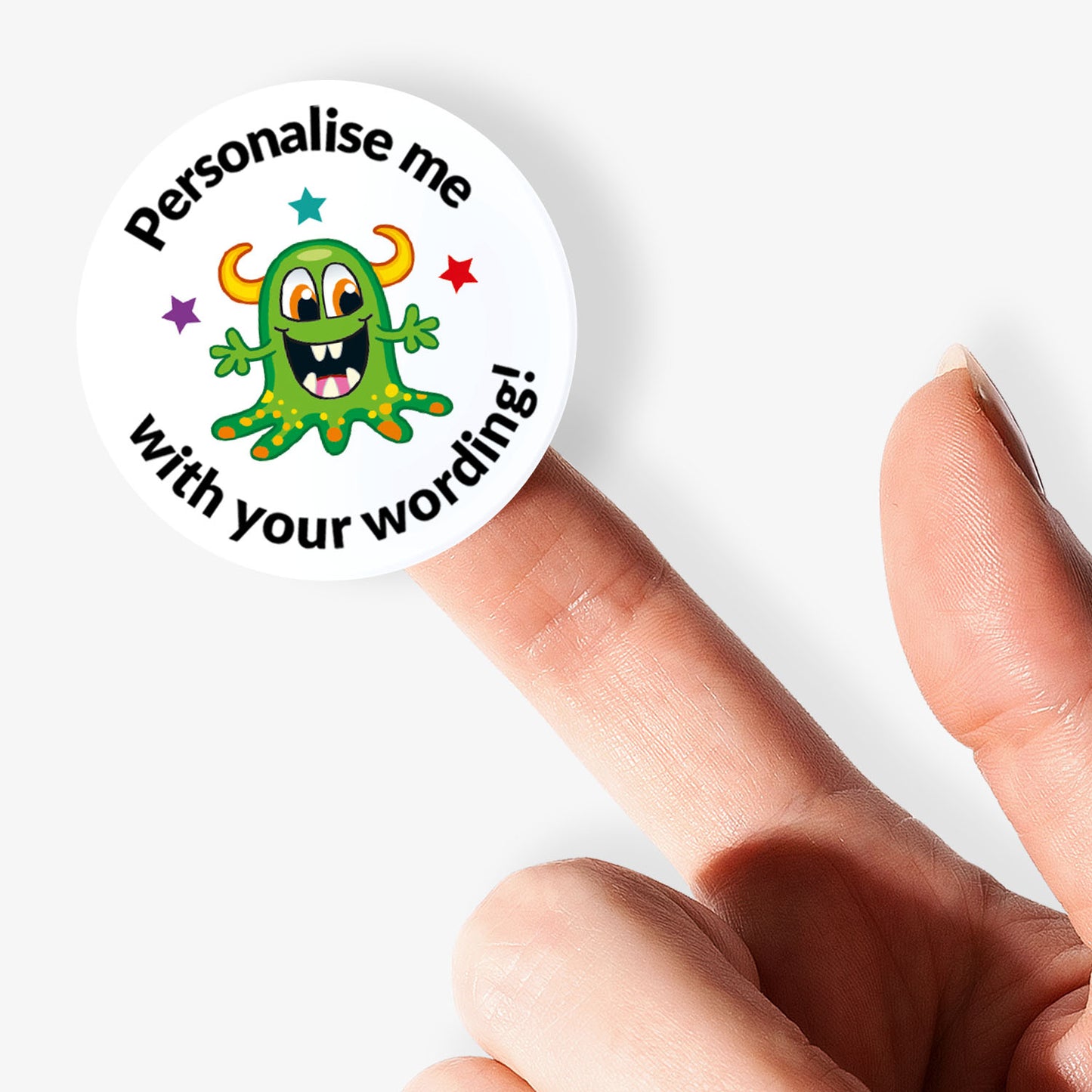 Circular sticker with a green monster graphic and text 'Personalise me with your wording!' held between fingers.