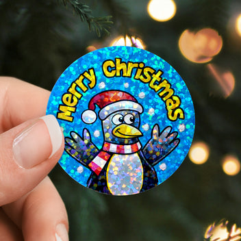 Circular Christmas sticker with a penguin design and 'Merry Christmas' text, held against a blurred Christmas tree background.