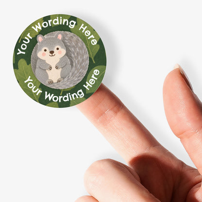 A close-up of a person's finger holding a circular custom sticker featuring a grey squirrel on a green leafy background with placeholder text 'Your Wording Here'.
