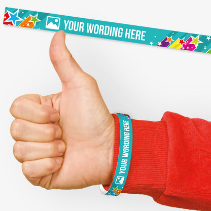 A customisable wristband with a cyan background with shooting stars of vibrant colours.  The centre is displaying the placeholder text 'YOUR WORDING HERE' alongside an 'choose image' icon. Also shown on a wrist of a person in a red jumper giving a thumbs up signal.