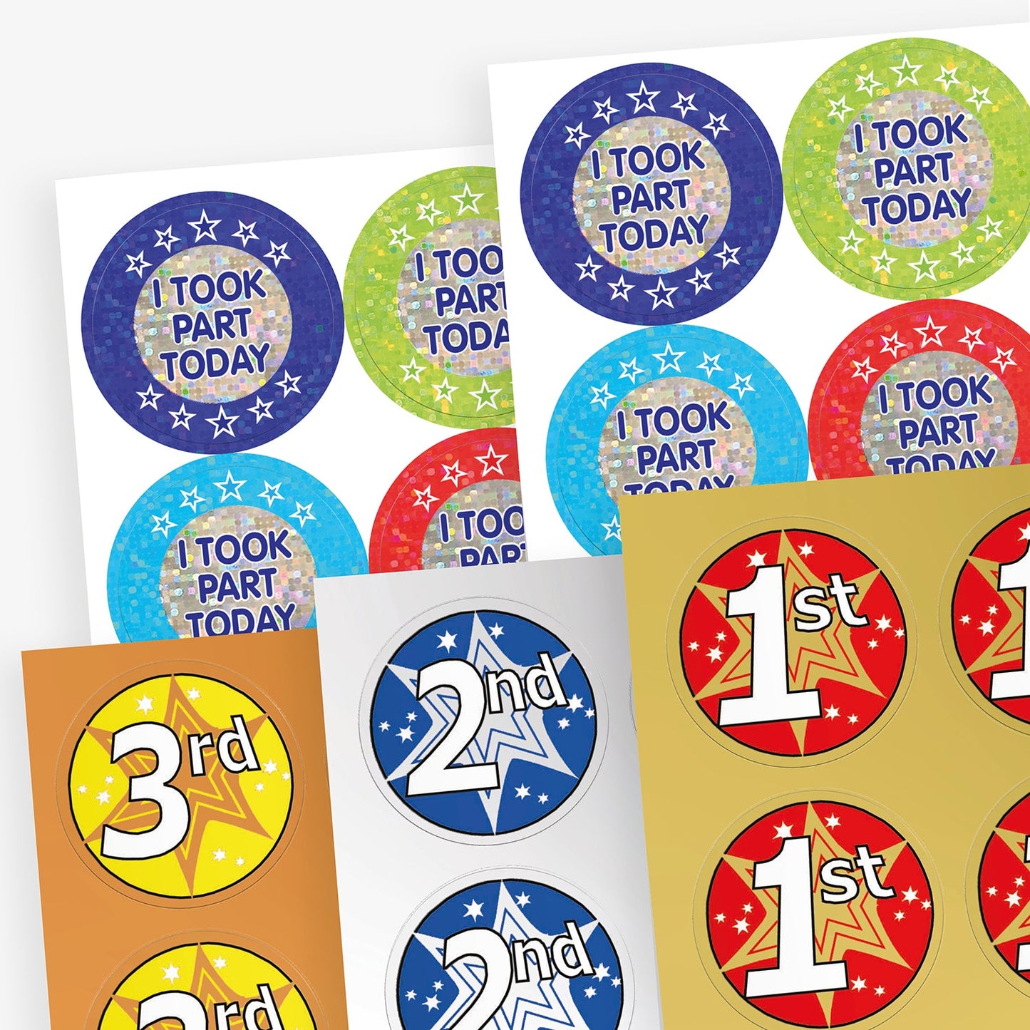A selection of circular holographic award stickers with phrases such as 'I Took Part Today', '1st', '2nd', and '3rd'. The 'I Took Part Today' stickers appear in various colours with star patterns, while the '1st', '2nd', and '3rd' place stickers feature bold numbers on red, silver, and bronze backgrounds with starburst designs.