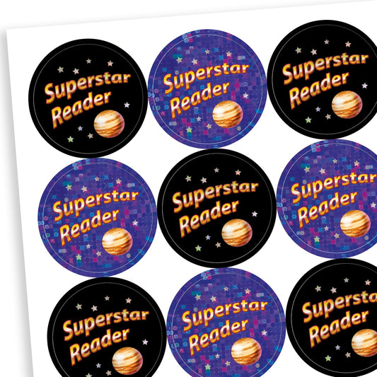 A sheet of circular, holographic reward stickers with the phrase 'Superstar Reader' in bright text. Each sticker features a planet illustration and a starry background. Half the stickers have a black background with orange and white text, while the other half have a sparkly purple background with blue and orange text, creating a cosmic theme.