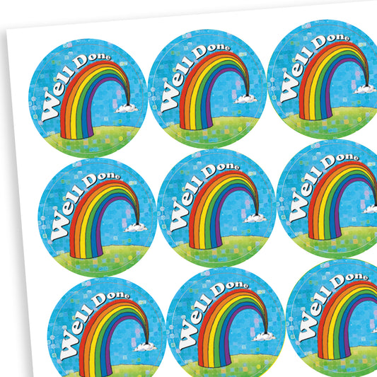 A sheet of holographic circular reward stickers. Each sticker features a colourful rainbow arcing from grassy hills toward small clouds, set against a bright blue sky with sparkly accents. The words 'Well Done' are written in bold white text along the curve of the rainbow.
