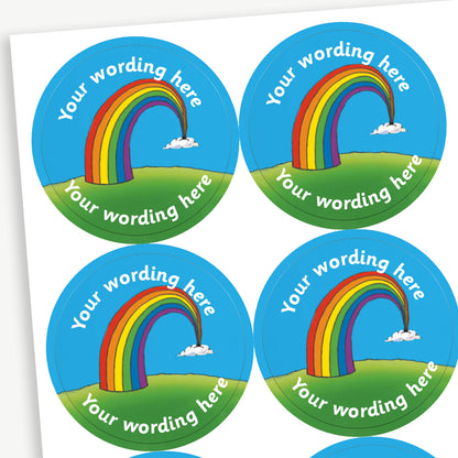 A sheet of circular reward stickers. Each sticker features a colourful rainbow arcing from grassy hills toward small clouds, set against a bright blue sky. Placeholder text 'Your Wording Here' in white font is above and below the rainbow for personalisation.