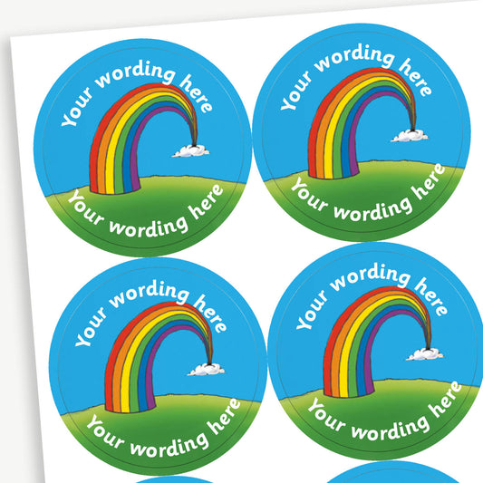 A sheet of circular reward stickers. Each sticker features a colourful rainbow arcing from grassy hills toward small clouds, set against a bright blue sky. Placeholder text 'Your Wording Here' in white font is above and below the rainbow for personalisation.