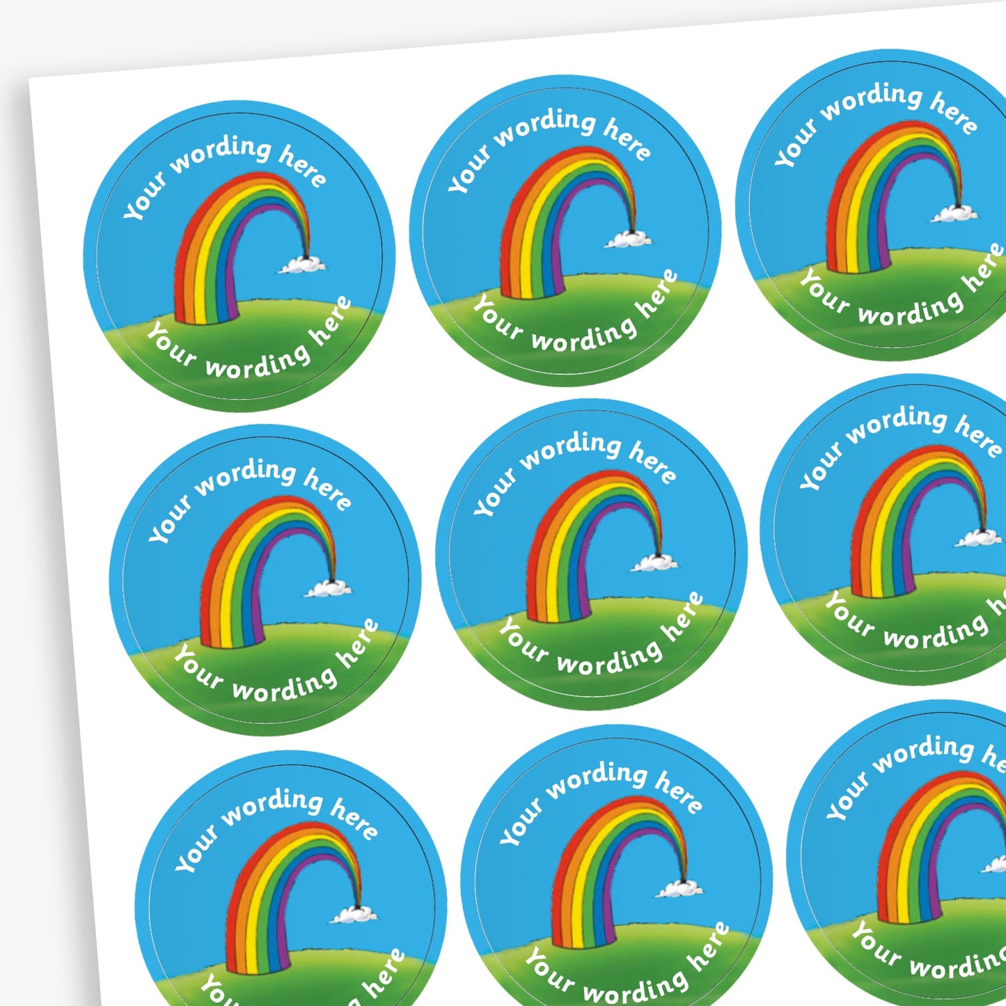 A sheet of circular reward stickers. Each sticker features a colourful rainbow arcing from grassy hills toward small clouds, set against a bright blue sky. Placeholder text 'Your Wording Here' in white font is above and below the rainbow for personalisation.