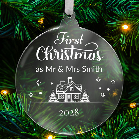 Round clear acrylic Christmas tree ornament with white design reading 'First Christmas as Mr & Mrs Smith 2028', featuring a house with trees, stars, and snowflake details, hanging on a green tree with fairy lights.