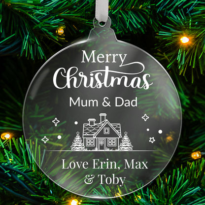Round clear acrylic Christmas tree ornament with white design reading 'Merry Christmas Mum & Dad', featuring a house with trees, stars, and snowflake details, with bottom customisable text 'Love Erin, Max & Toby' hanging on a green tree with fairy lights.