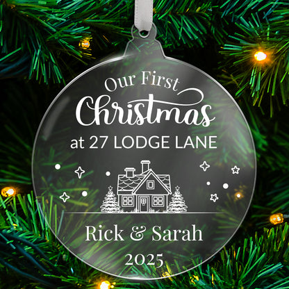 Round clear acrylic Christmas tree ornament with white design reading 'Our First Christmas at 27 LODGE LANE', featuring a house with trees, stars, and snowflake details, with bottom customisable text 'Rick & Sarah 2025' hanging on a green tree with fairy lights.