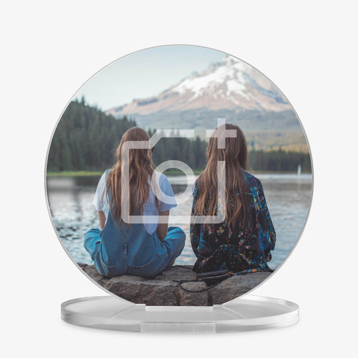 A round acrylic photo display on a clear stand, featuring an image of two girls sitting on a stone edge facing a serene lake with a snow-capped mountain in the distance. One girl is wearing blue overalls and a white shirt, and the other is in a dark floral outfit. A faint camera icon overlay indicates the area is customisable with a personal photo.