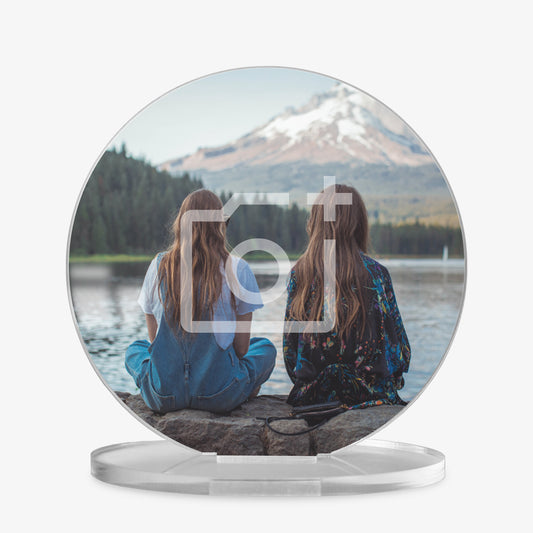 A round acrylic photo display on a clear stand, featuring an image of two girls sitting on a stone edge facing a serene lake with a snow-capped mountain in the distance. One girl is wearing blue overalls and a white shirt, and the other is in a dark floral outfit. A faint camera icon overlay indicates the area is customisable with a personal photo.