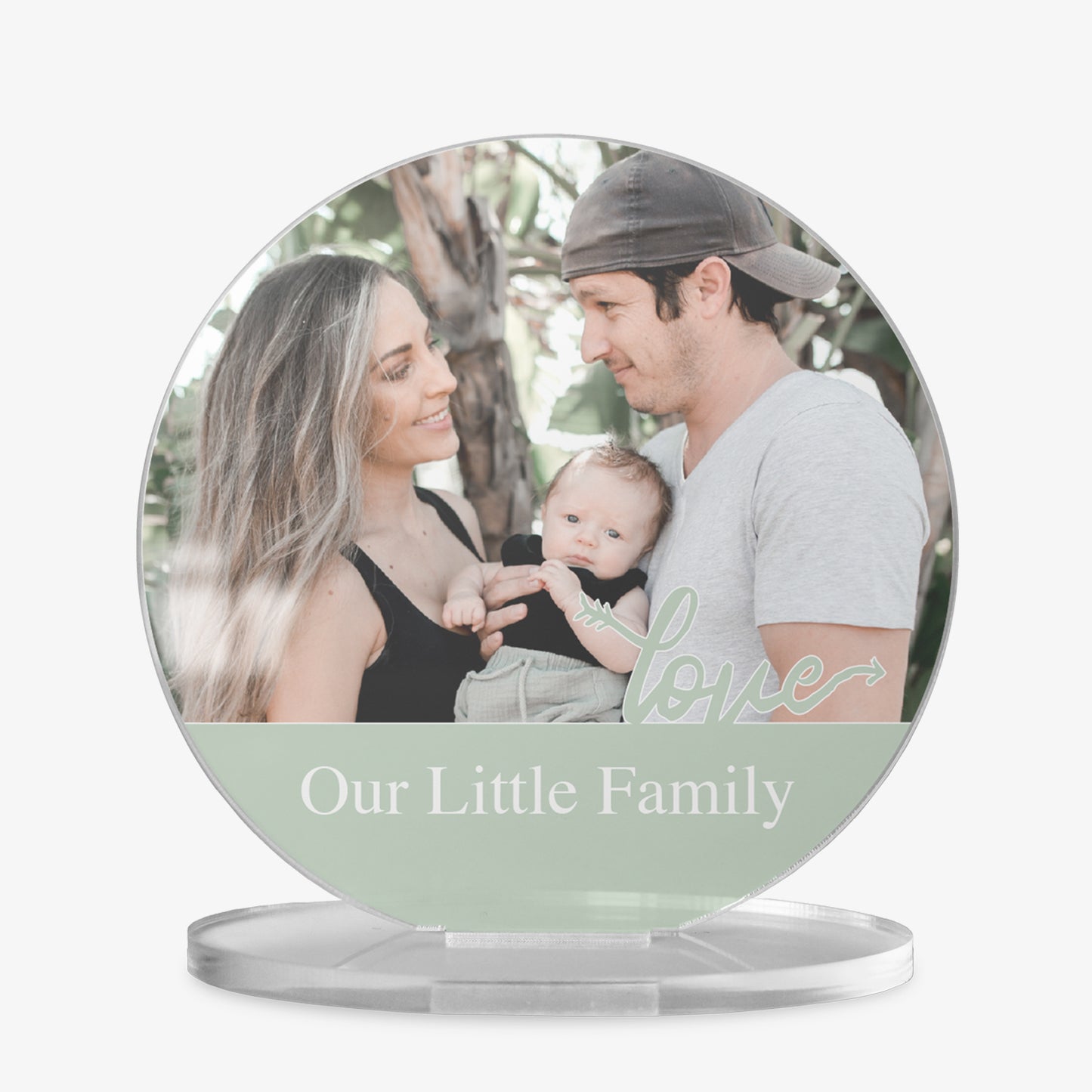 A round acrylic display piece with a couple holding their baby with trees in the background. The bottom section has a sage band with the word 'love' in cursive and space for personalised text reading 'Our Little Family'. The item is displayed on a clear round stand.