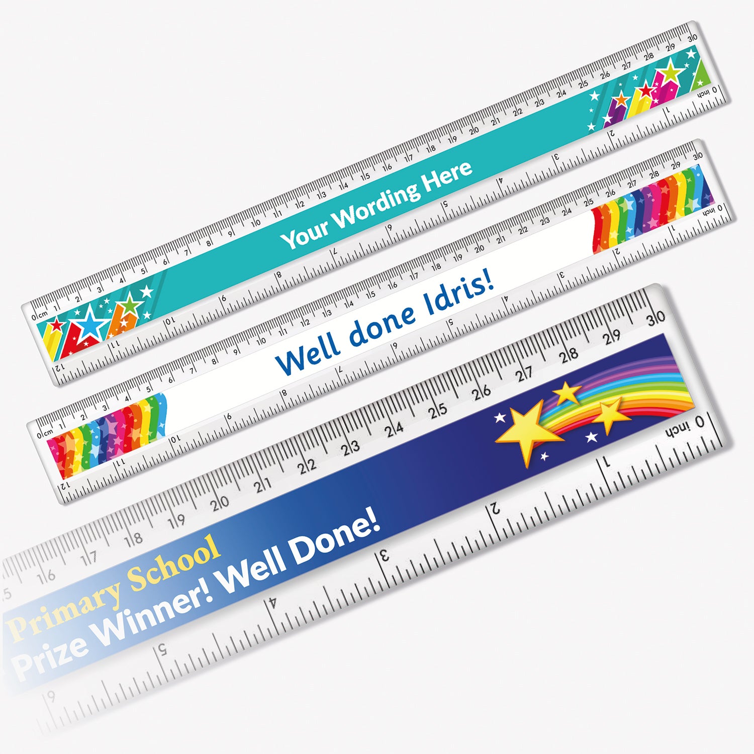 3 colourful 30cm plastic rulers, each with a printed design and measurement markings in cm and mm. The top ruler has a turquoise background with rainbow letters and the phrase 'Your Wording Here'. The middle ruler has a white background with rainbow pencils at each end and text 'Well done Idris!' in blue. The bottom ruler features a navy blue background with shooting stars and the text 'Primary School Prize Winner! Well Done!' in bold white lettering.