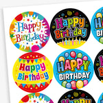 A sheet of circular 'Happy Birthday' stickers featuring various vibrant designs. Each sticker includes colourful text and festive decorations such as balloons, candles, stars, and bunting, with background colours including black, blue, yellow, and multicolour.