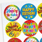 A sheet of colourful, holographic circular 'Happy Birthday' stickers with celebratory designs. Each sticker features bright backgrounds in blue, yellow, and red, decorated with confetti, balloons, and streamers, with bold 'Happy Birthday' text in various styles.