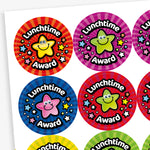 A sheet of brightly coloured circular stickers with the phrase 'Lunchtime Award' in bold white text on a black curved banner. Each sticker features a smiling cartoon star character in the centre, surrounded by colourful stars. Background colours include red, purple, blue, yellow, and green, with vibrant radial patterns that create a cheerful and eye-catching effect.