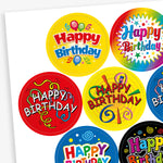 Sheet of strawberry scented round birthday stickers in various bright colours and designs. Each sticker features the phrase 'Happy Birthday' in different playful fonts, with decorative elements such as balloons, stars, swirls, and confetti. Backgrounds include red, yellow, rainbow, blue, and black themes.
