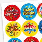 Sheet of strawberry scented round birthday stickers in various bright colours and designs. Each sticker features the phrase 'Happy Birthday' in different playful fonts, with decorative elements such as balloons, stars, swirls, and confetti. Backgrounds include red, yellow, rainbow, blue, and black themes.
